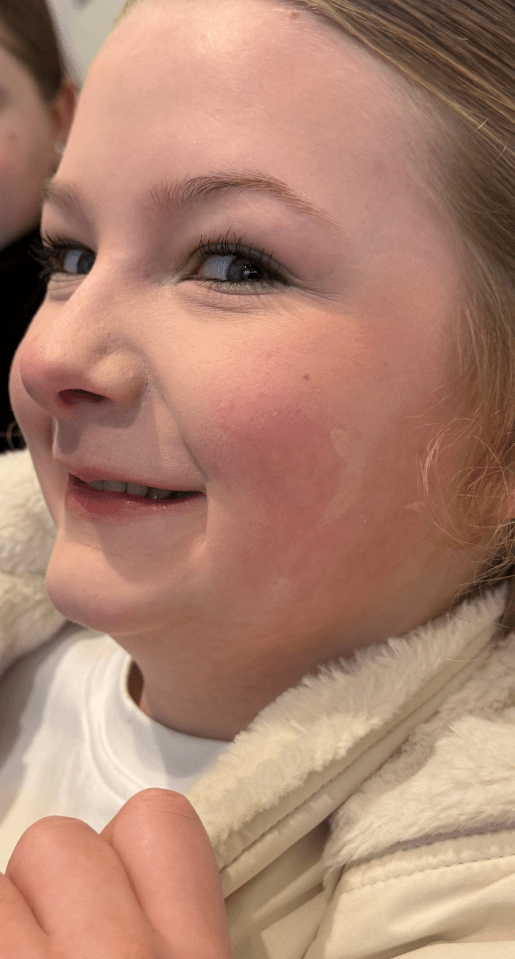 A young girl with blonde hair smiling while wearing makeup, including eyeliner and blush.