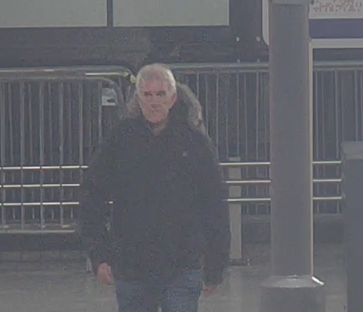 A man with gray hair and a dark jacket walks past a metal fence.