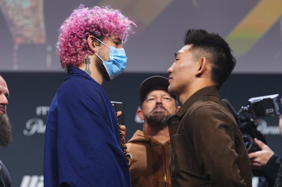 A masked fighter with pink hair and a blue cloak faces an unmasked fighter in a brown jacket at a UFC press conference.