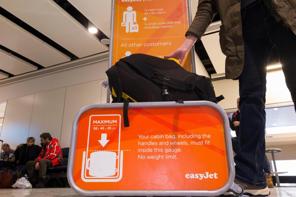 A person placing a black backpack into an EasyJet bag gauge at London Gatwick Airport.