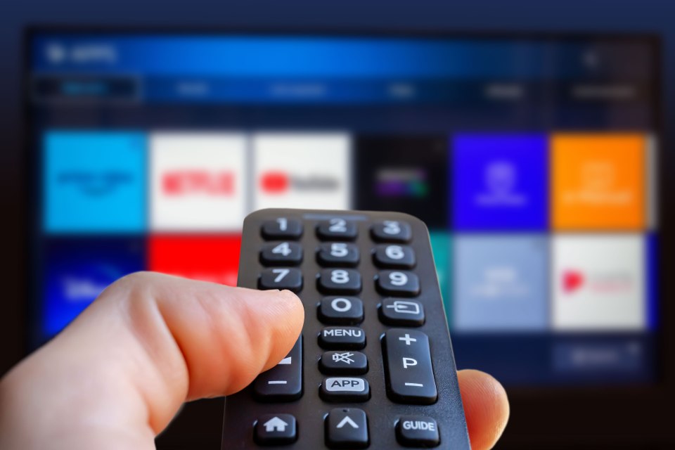 A person holding a smart TV remote control, with a blurred TV screen showing streaming apps in the background.