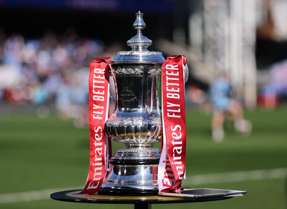 FA Cup trophy with red ribbons on a table on a soccer field.