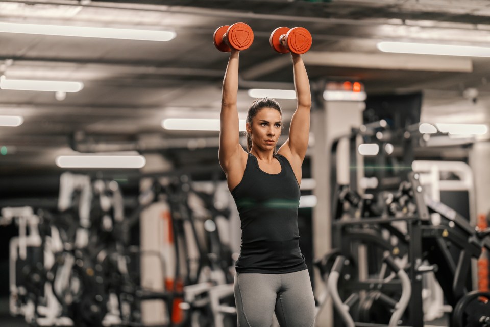 Mid-adult woman performing overhead dumbbell presses in a gym.
