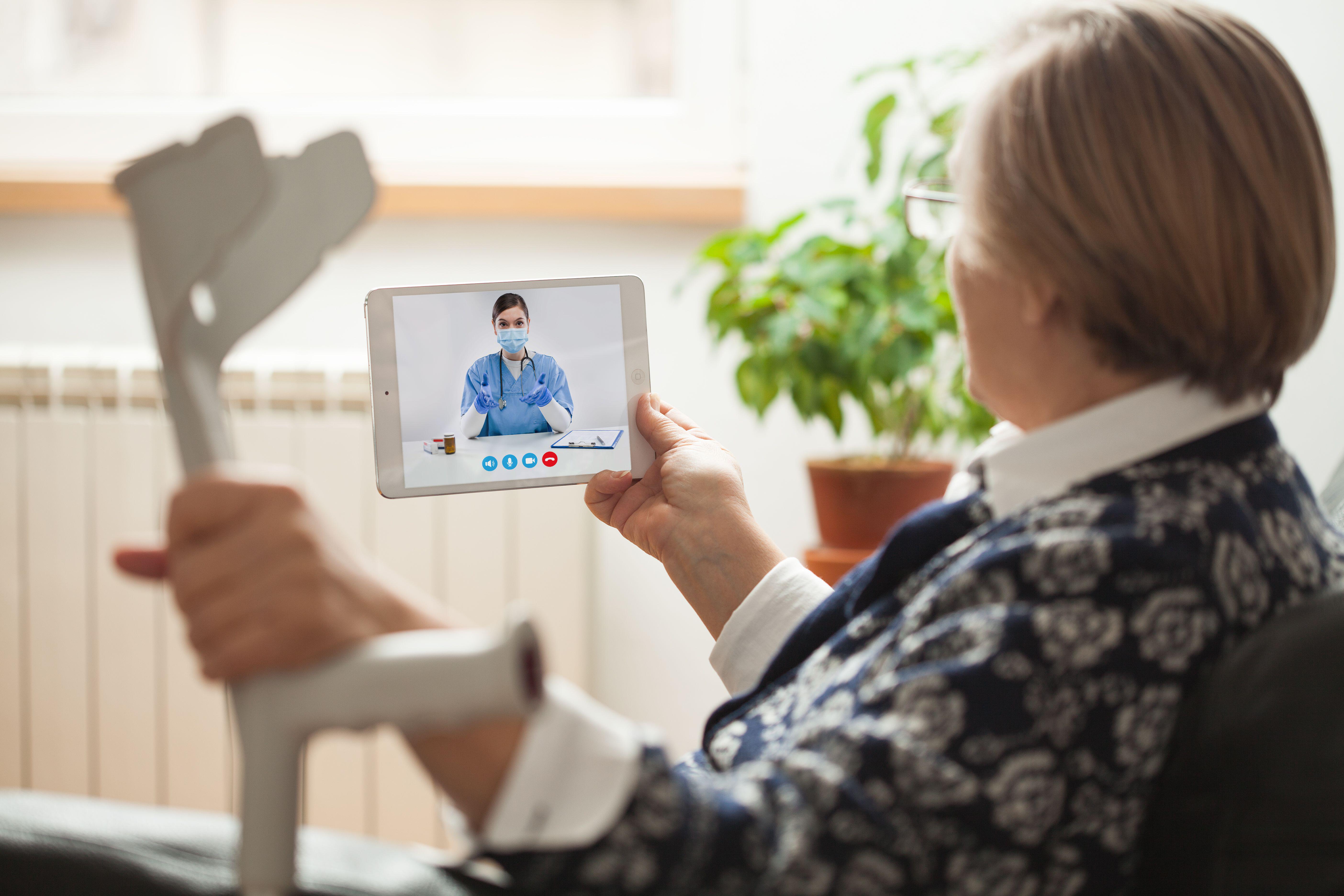 Retired senior elderly woman holding crutch and tablet computer,talking to NHS GP orthopedic female doctor via virtual telemedicine video call,broken