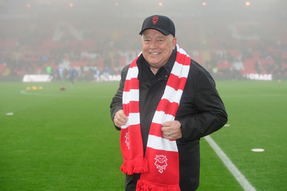 Ron Fowler, director of Lincoln City, smiling in a black jacket, cap, and red and white striped scarf on a football pitch.