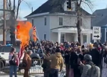 A man in a tan jacket was seen waving a burning upside down American flag in front of a large group of protesters on Wednesday, just hours after a 37-year-old woman was shot dead by ICE