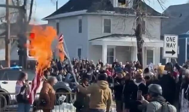 A man in a tan jacket was seen waving a burning upside down American flag in front of a large group of protesters on Wednesday, just hours after a 37-year-old woman was shot dead by ICE