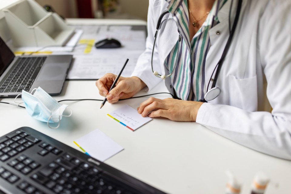 A female doctor, wearing a lab coat and stethoscope, making notes while on a video call.