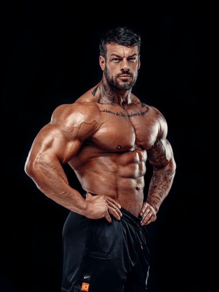 Jamie Christian Johal, known as Giant, a bodybuilder with visible tattoos, wearing black shorts, standing with hands on hips.