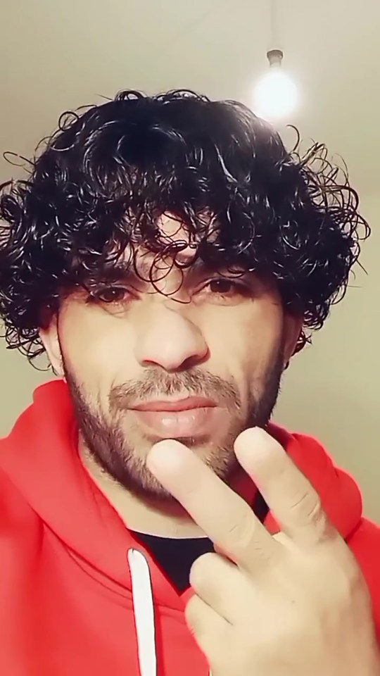 A man with dark curly hair, beard, and red hoodie holds up a peace sign.