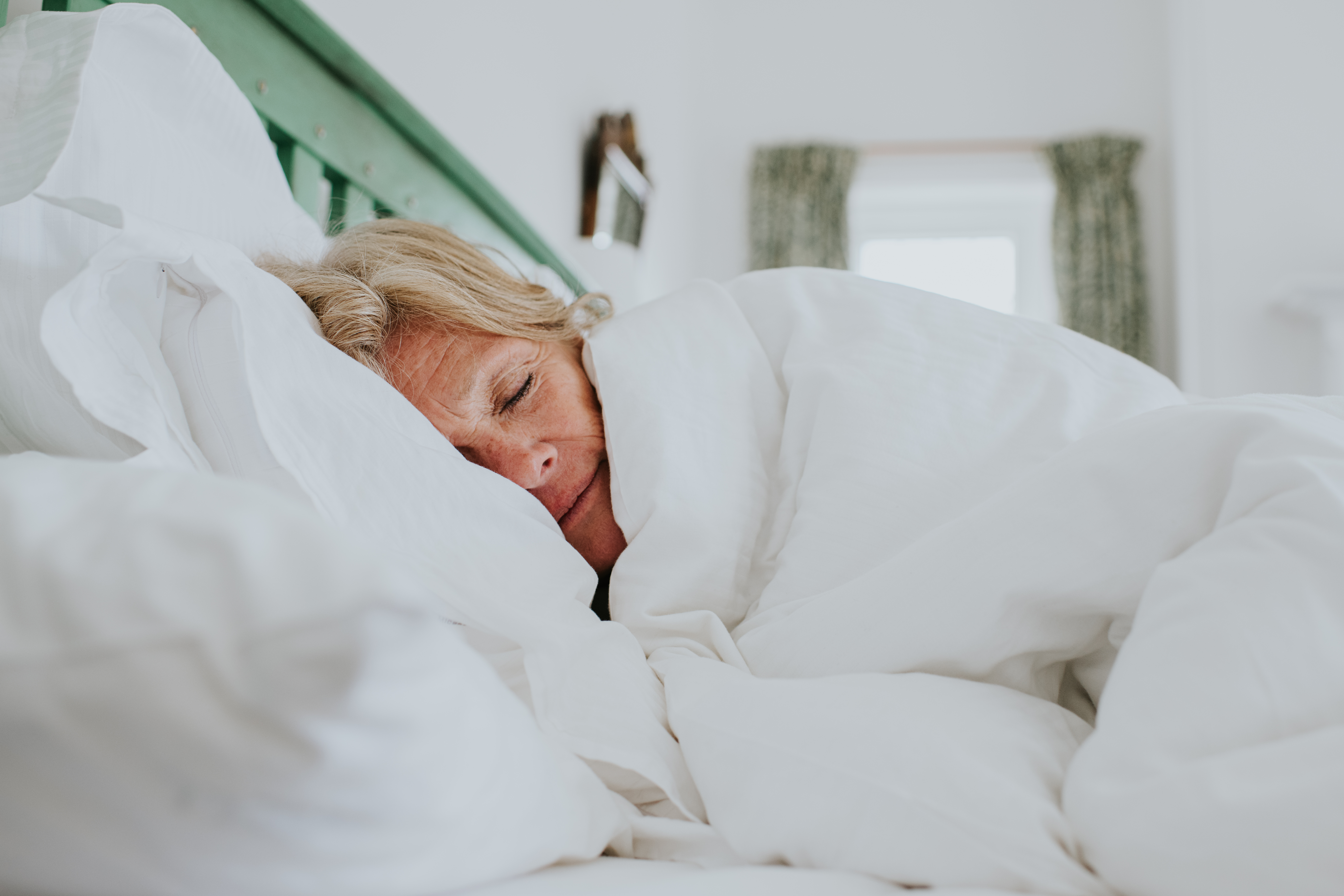 A mature woman snuggles up in a clean, white duvet in a bright and airy bedroom