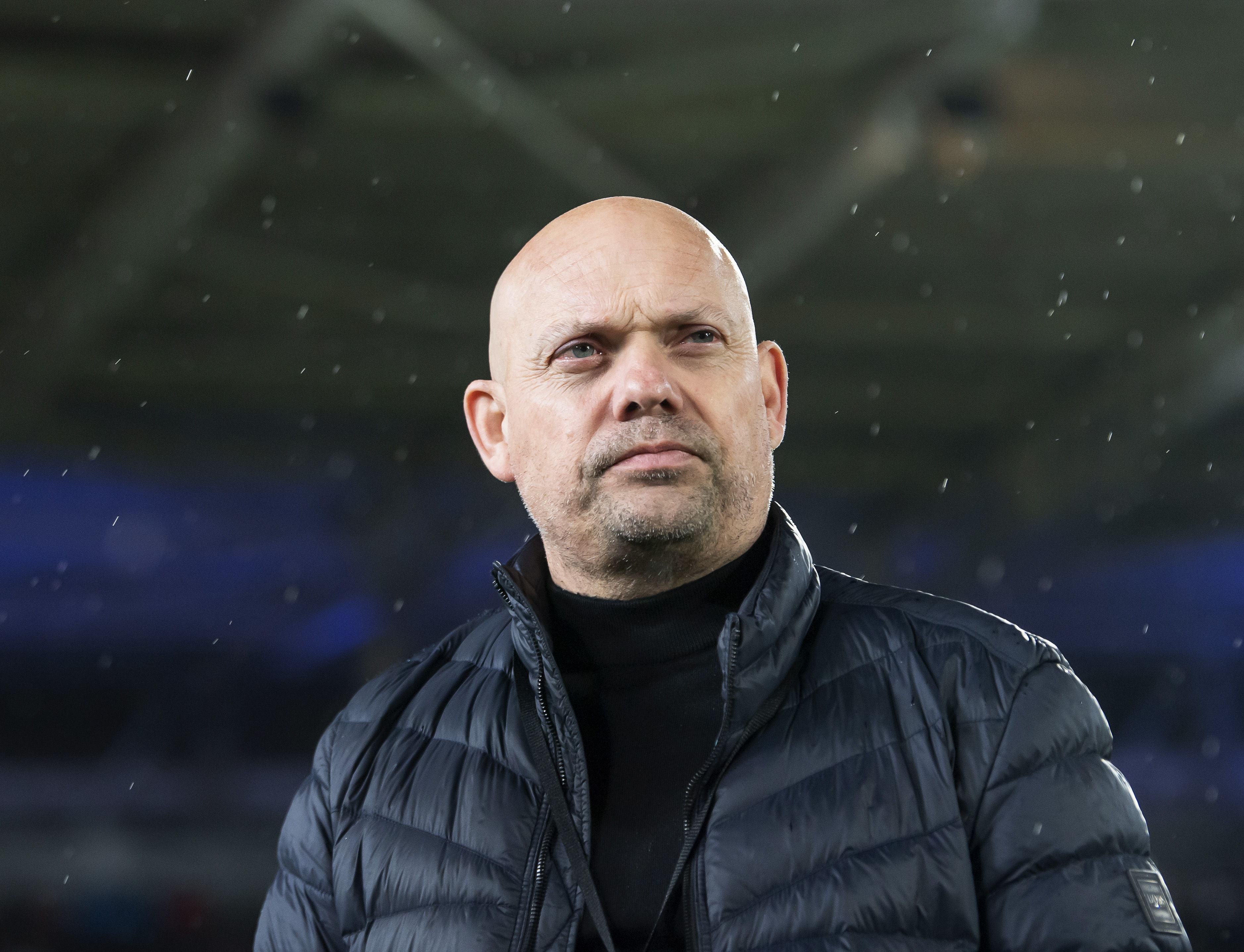 A bald man in a dark puffer jacket and black turtleneck looks off to the side, with blurry rain in the foreground.