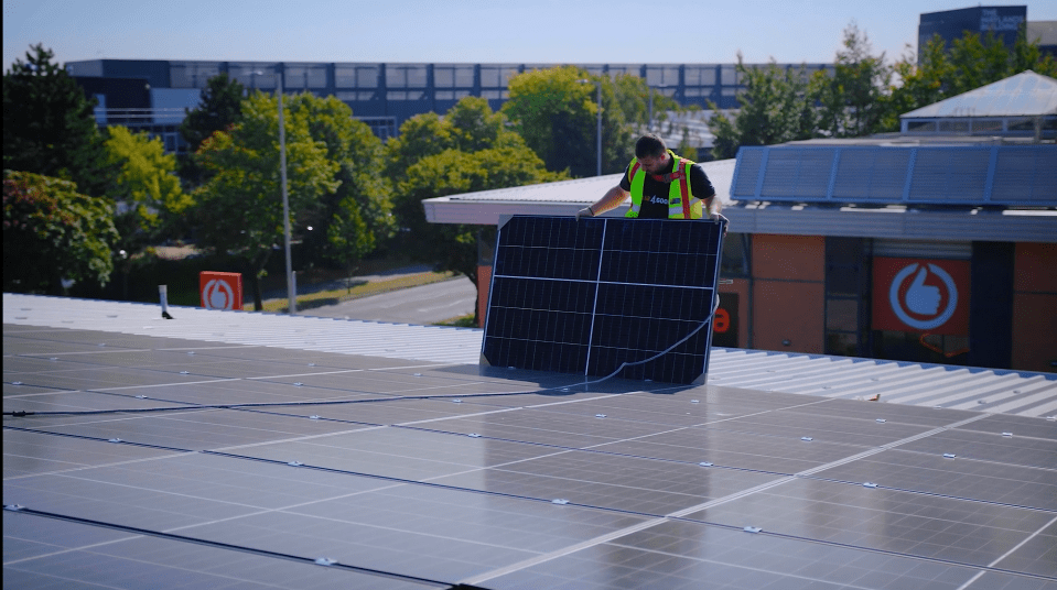 A person in a safety vest installs a solar panel on a rooftop with other solar panels.