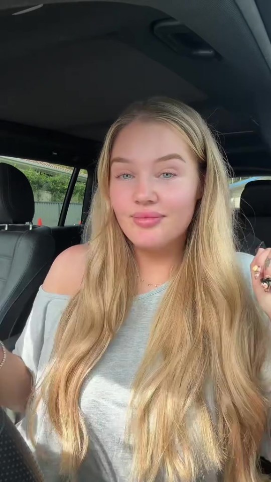 A woman with long blonde hair and light blue eyes looks at the camera while sitting in a car.