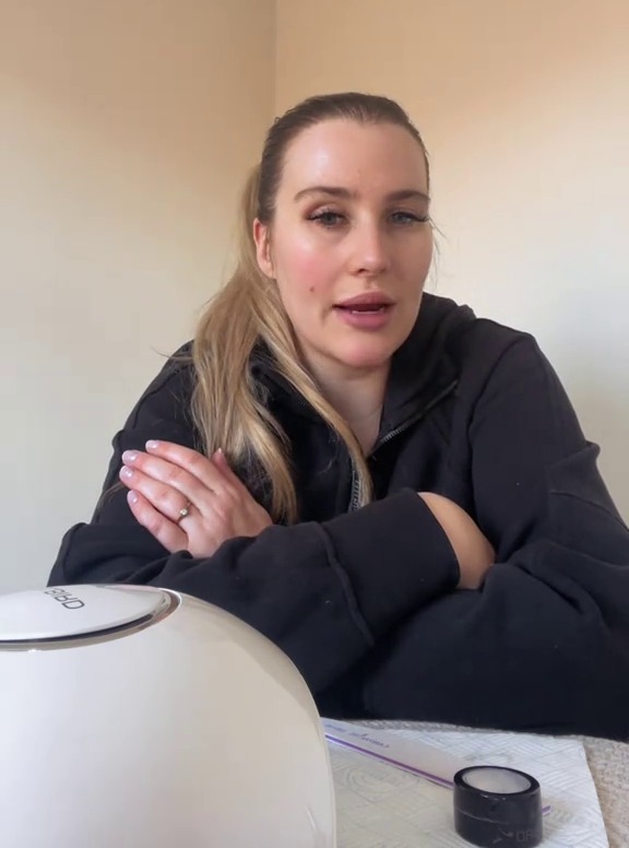 A woman with long blonde hair and wearing a black hoodie talks to the camera, with a nail UV lamp visible in the foreground.