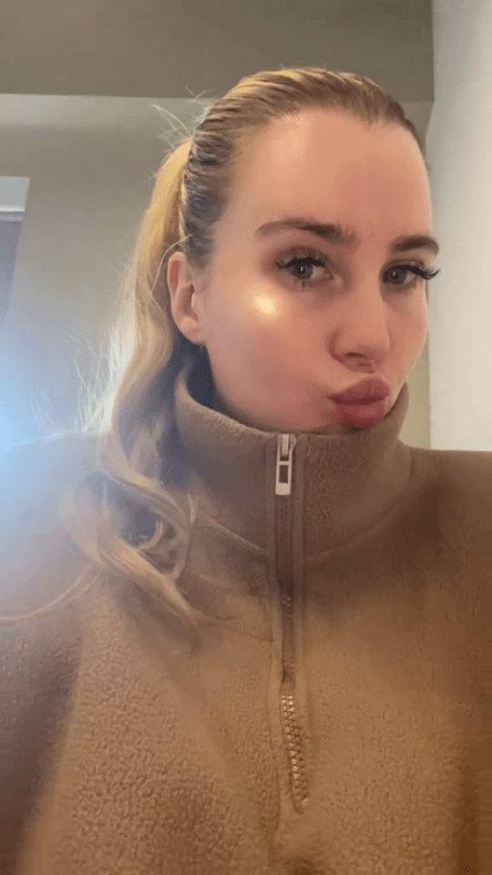 A woman with blonde hair in a ponytail wearing a brown fleece half-zip top, pouting at the camera.