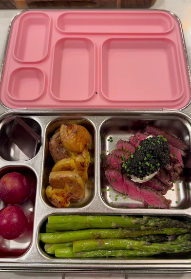 A lunchbox filled with sliced steak topped with caviar, roasted potatoes, asparagus, plums, and a chocolate bar.