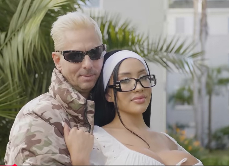 A man with bleached blonde hair and sunglasses embraces a woman wearing a white headband and large-framed glasses.