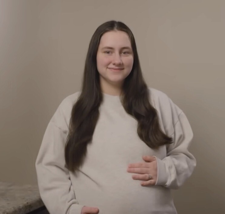 A young woman with long dark hair, wearing a light-colored sweatshirt, with one hand resting on her pregnant belly.
