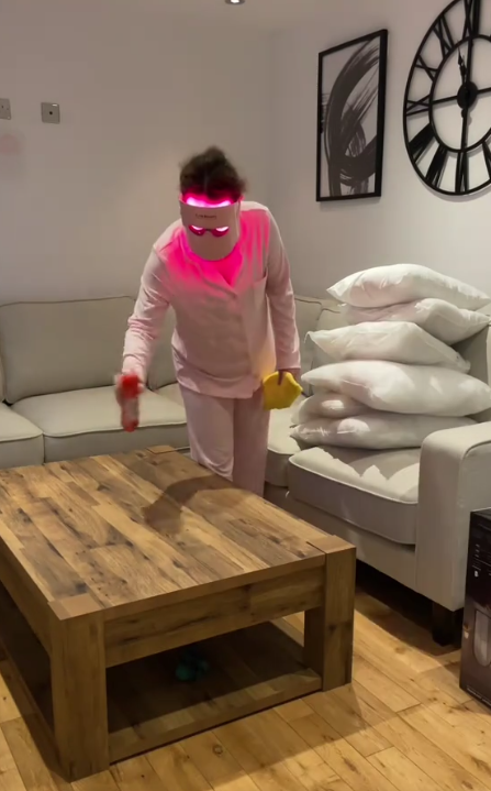 A person in pink pajamas and a bright pink light-up visor cleans a coffee table with a spray bottle and cloth.