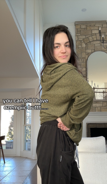 A woman in a green hoodie and black sweatpants stands with her back partially turned, looking over her shoulder with a smirk, with text reading "you can tell I have ozempiic buttt....."