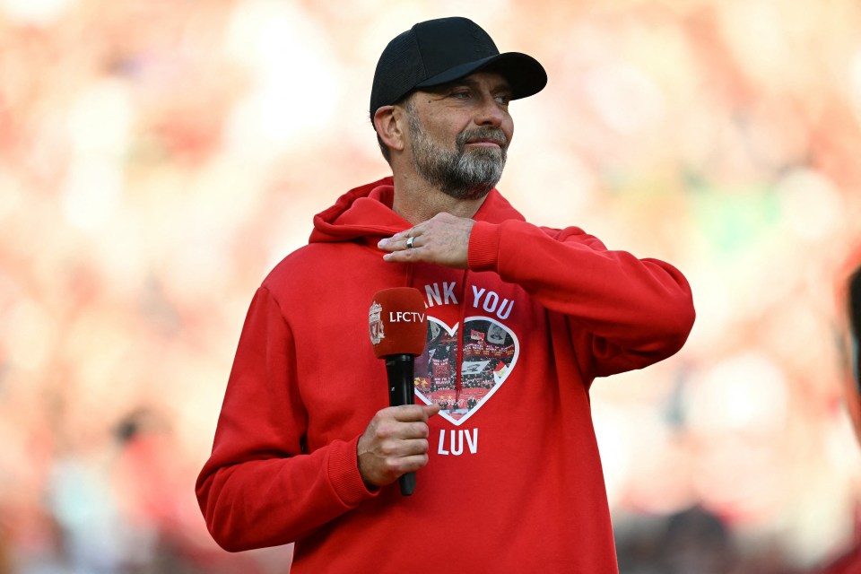 Liverpool manager Jurgen Klopp holds a microphone and gestures with his other hand on his chest, wearing a red hoodie with a "Thank You" heart design.