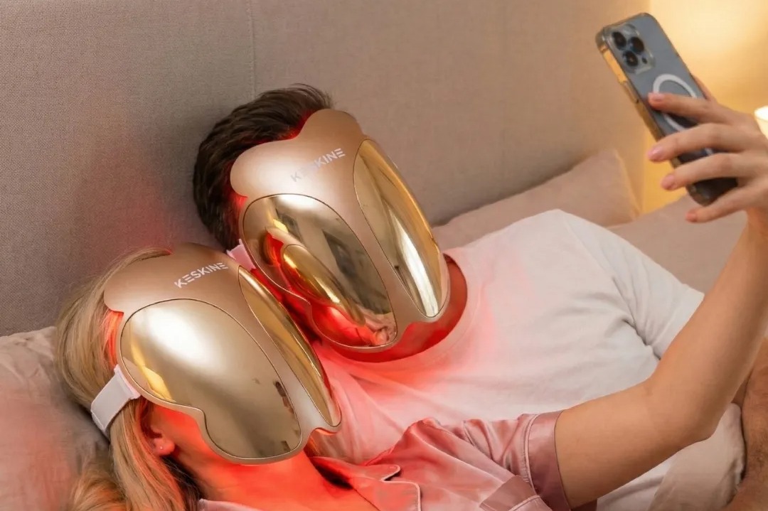 Two people wearing gold LED face masks, with the person in front holding a smartphone.