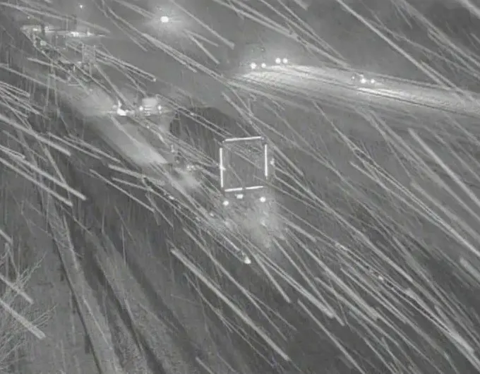 A highway camera view of a snowy road at night, with light trails from vehicle headlights and snow falling across the frame.