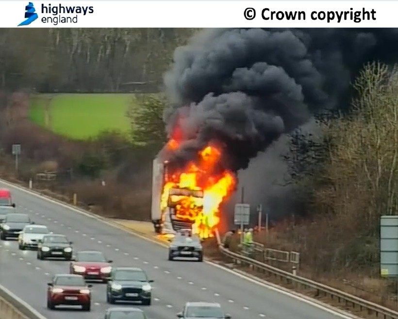 A truck engulfed in flames on the side of a multi-lane highway, with black smoke billowing into the sky.