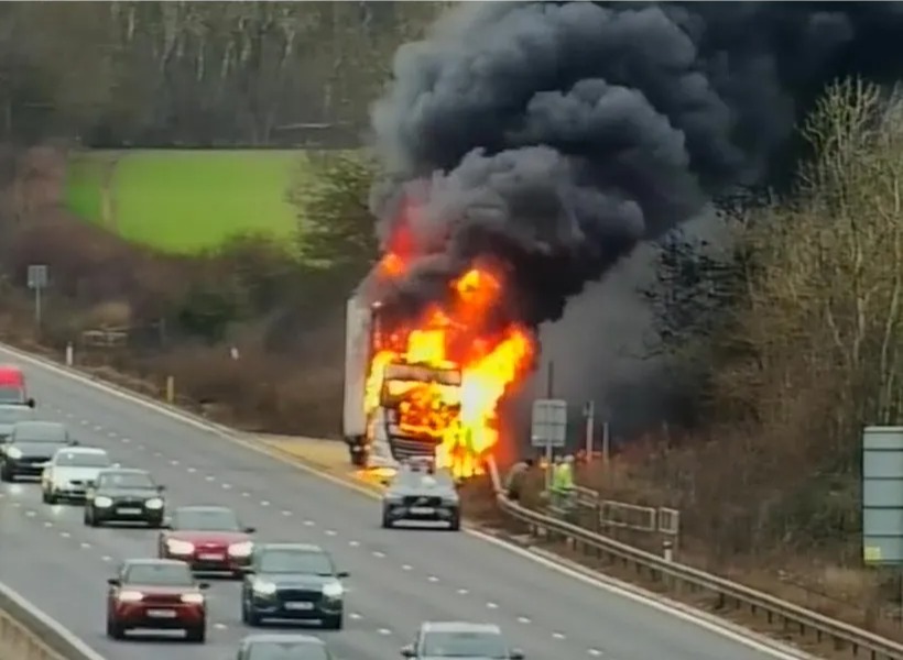 A truck engulfed in flames on the side of a multi-lane highway, with black smoke billowing into the sky.