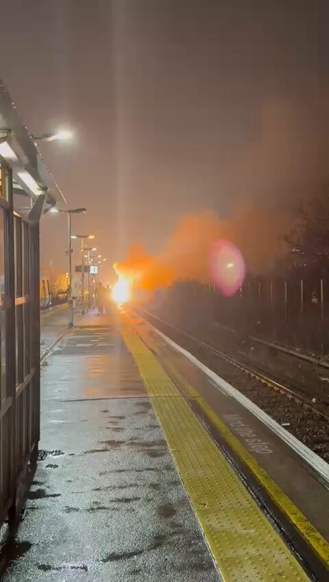 Large fire burning on train tracks at a station platform at night.