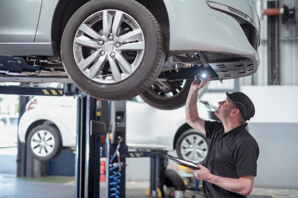 A mechanic uses a flashlight to inspect the underside of a car raised on a lift.