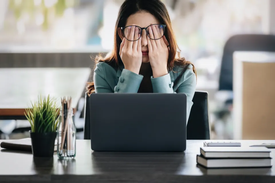 Woman wearing glasses rubbing her eyes while sitting at a desk with a laptop.