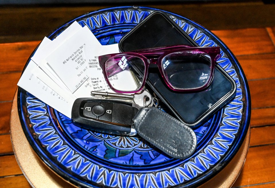 A set of car keys, a smartphone, magenta-rimmed glasses, and several paper receipts sitting in a blue and black decorative dish.