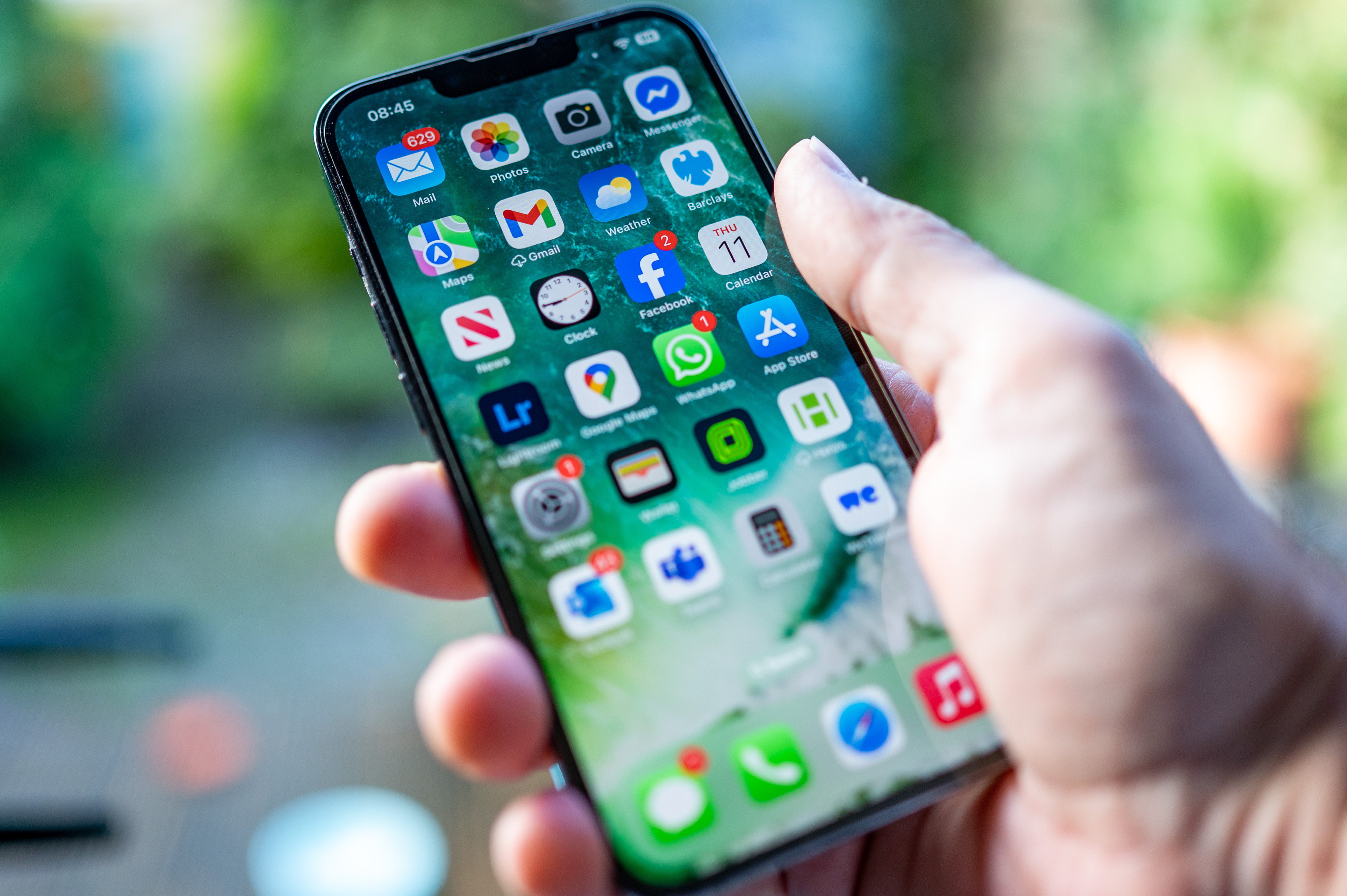 A hand holds an iPhone displaying various app icons on the home screen, including Mail, Photos, Camera, Messenger, Maps, Gmail, Weather, Facebook, Calendar, News, Clock, WhatsApp, App Store, and other apps.