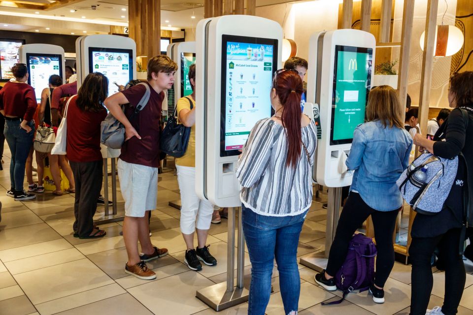 Crowds of people, including children, teenagers, and adults, gathered around self-service kiosks at a busy McDonald's restaurant in London's Leicester Square.