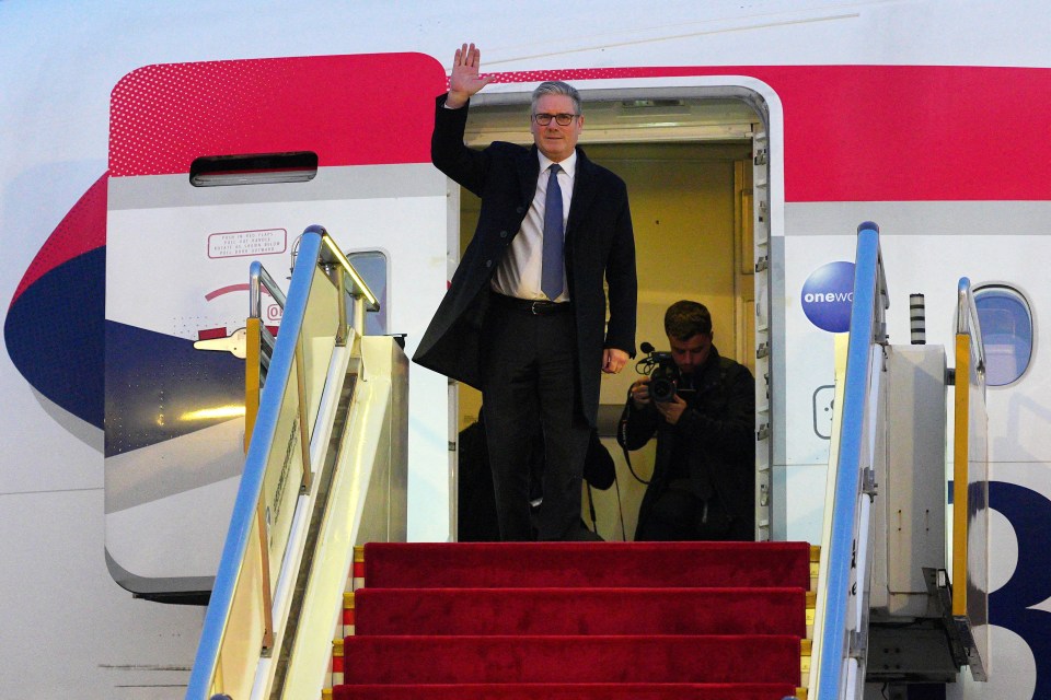 British Prime Minister Keir Starmer waves from the top of an airplane's red-carpeted stairs.