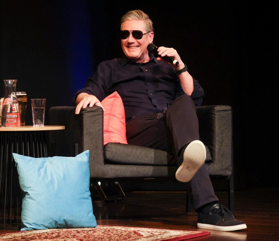 Keir Starmer, wearing sunglasses, sits relaxed in a chair with a microphone in hand.