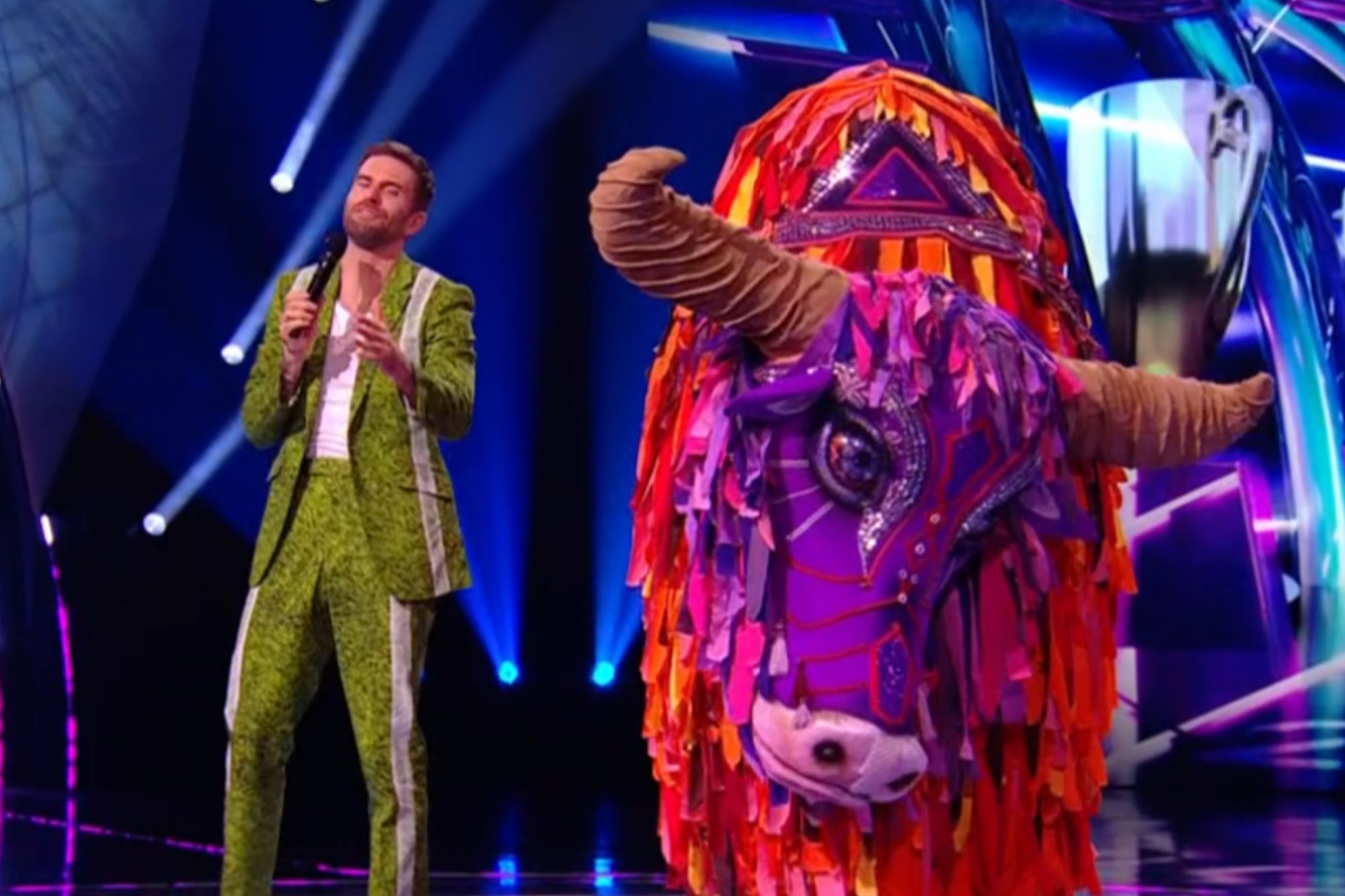An image collage containing 1 images, Image 1 shows A man in a green suit holding a microphone standing next to a large colorful yak costume on a stage