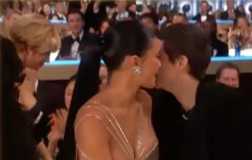 A woman in a sparkly dress leans in to kiss a man in a black shirt, while another person in a black suit watches them.
