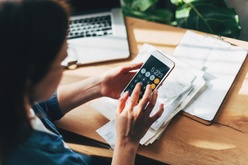 Person using a smartphone calculator to manage finances, with paper documents and a laptop nearby on a wooden desk.