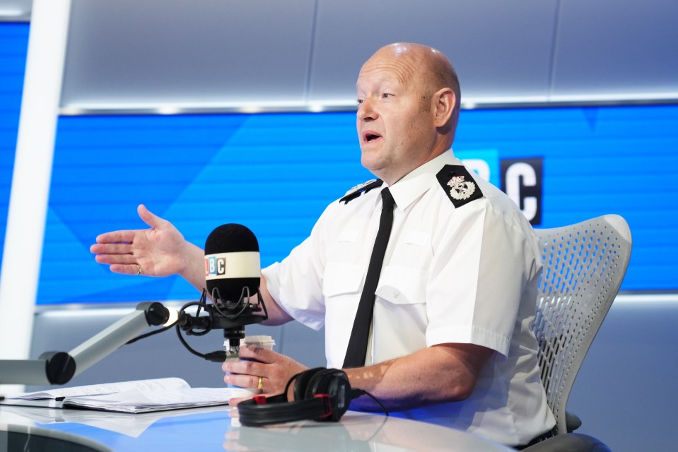 Chief Constable Craig Guildford speaks into a microphone at LBC Radio, gesturing with his left hand.