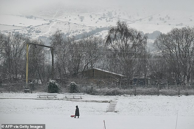 A pedestrian walks their dog across a snow-covered cricket pitch today in Glossop, Derbyshire
