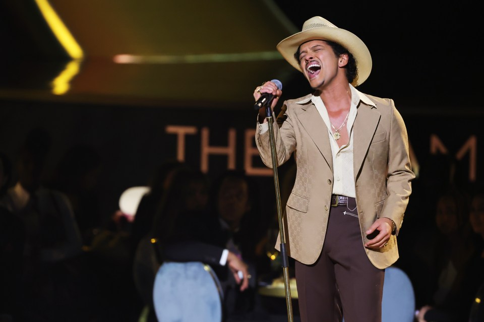 Bruno Mars singing into a microphone, wearing a tan cowboy hat, tan suit jacket, and brown pants, on stage at the 67th Annual GRAMMY Awards.