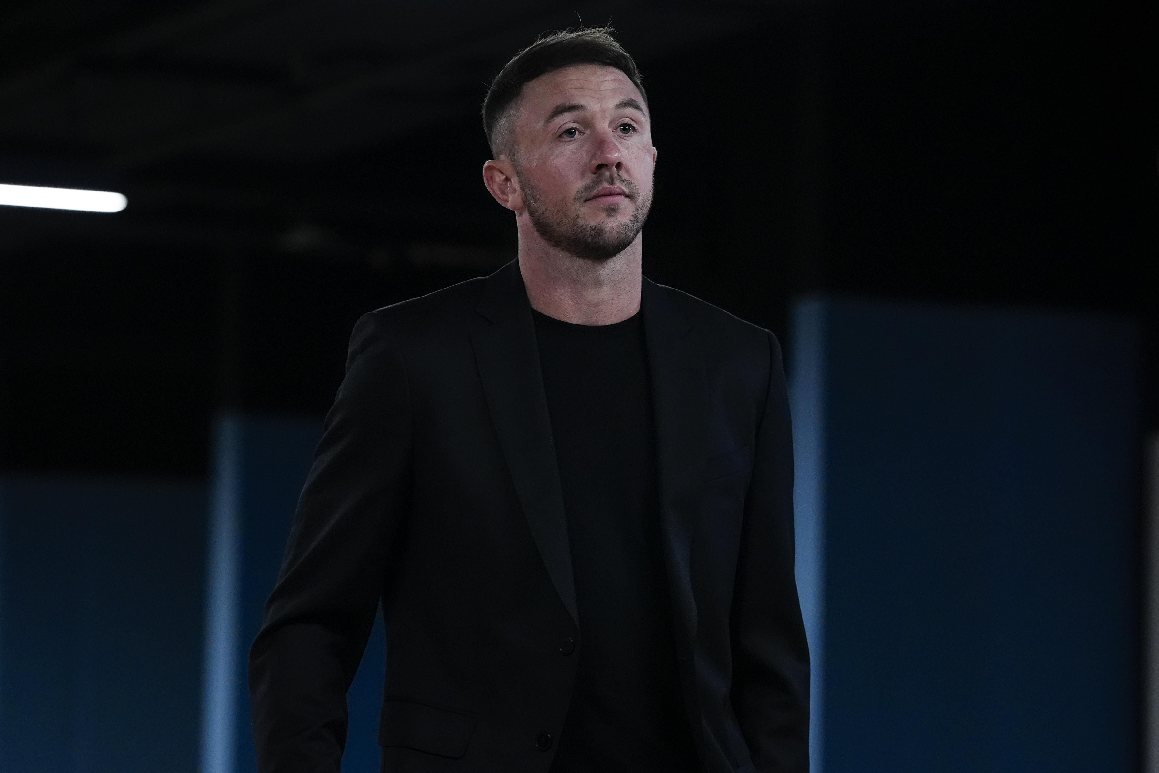 A man in a black jacket and t-shirt with short hair and a beard, looking off to the side.