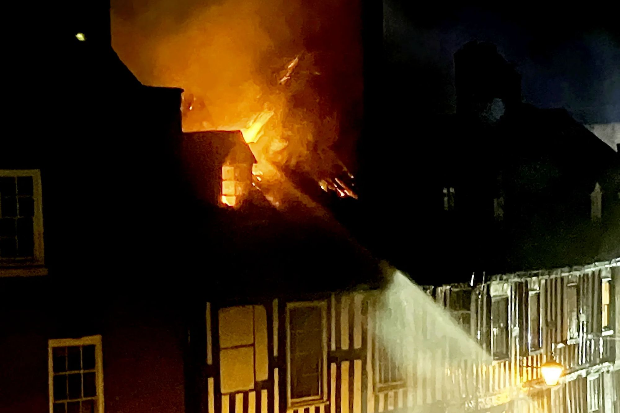 An image collage containing 1 images, Image 1 shows Firefighters spray water on a fire blazing from the roof of a historic timber-framed building at night