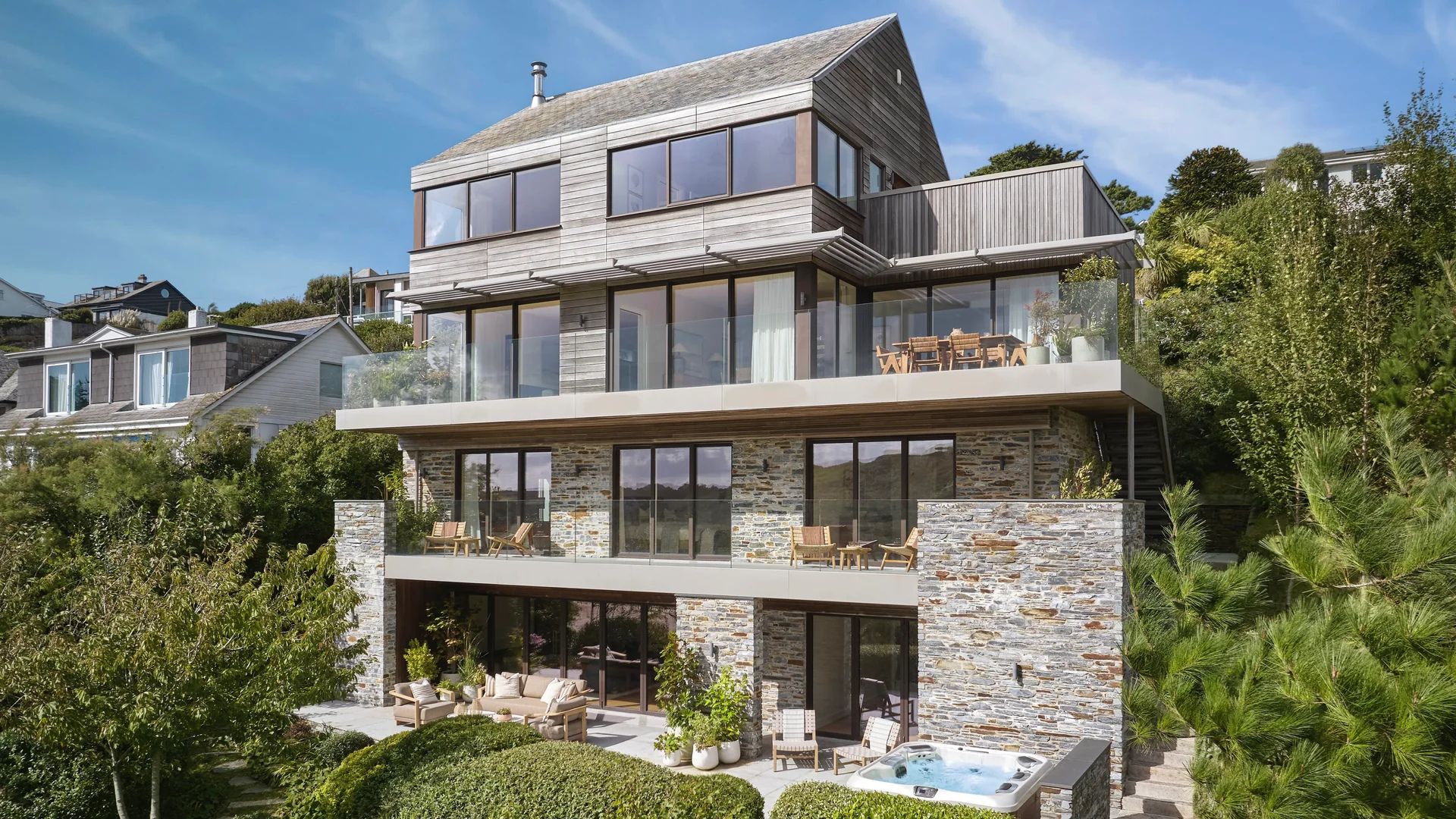 An aerial photo of a three-story house with natural wood siding and stone walls on a hillside, featuring multiple balconies and outdoor living spaces, and a hot tub.