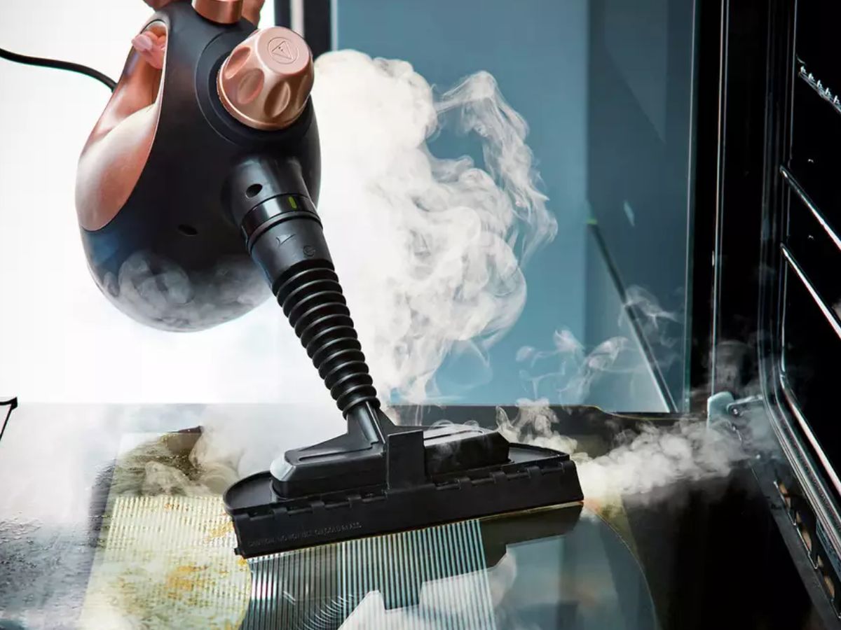 Handheld steam cleaner cleaning a grimy oven door.