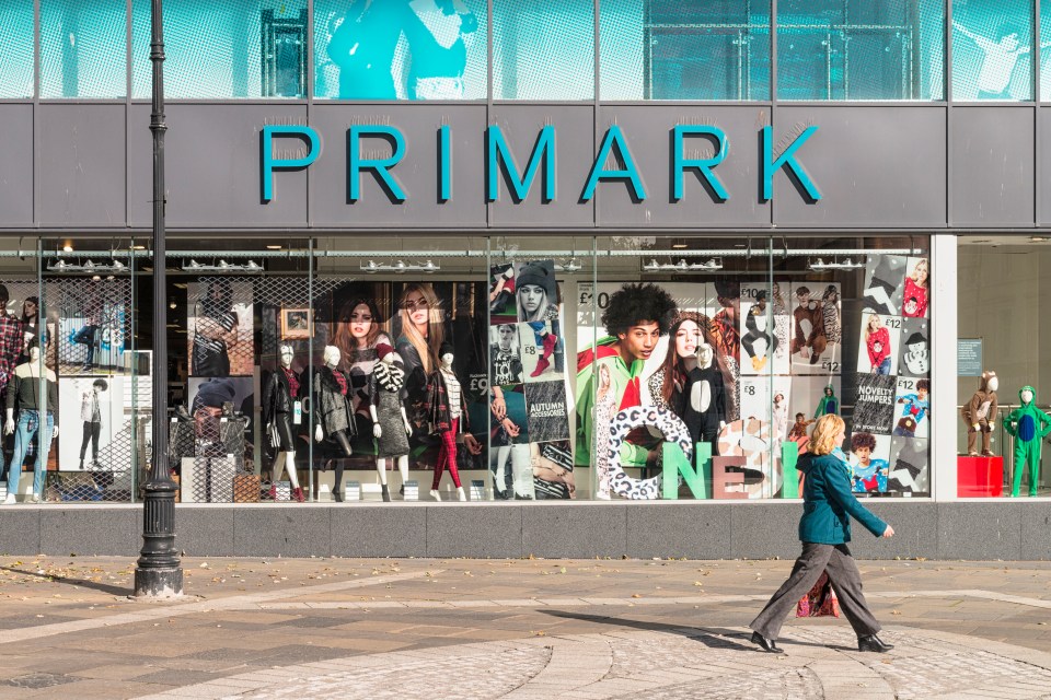 A woman walks past a Primark store window display featuring clothing and advertising.