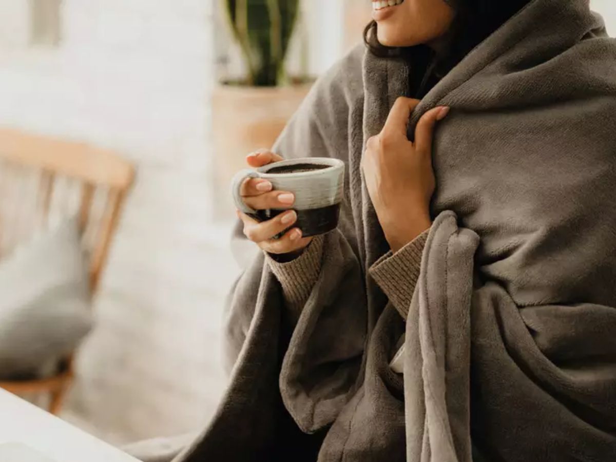 Person wrapped in a grey blanket holding a mug of coffee.